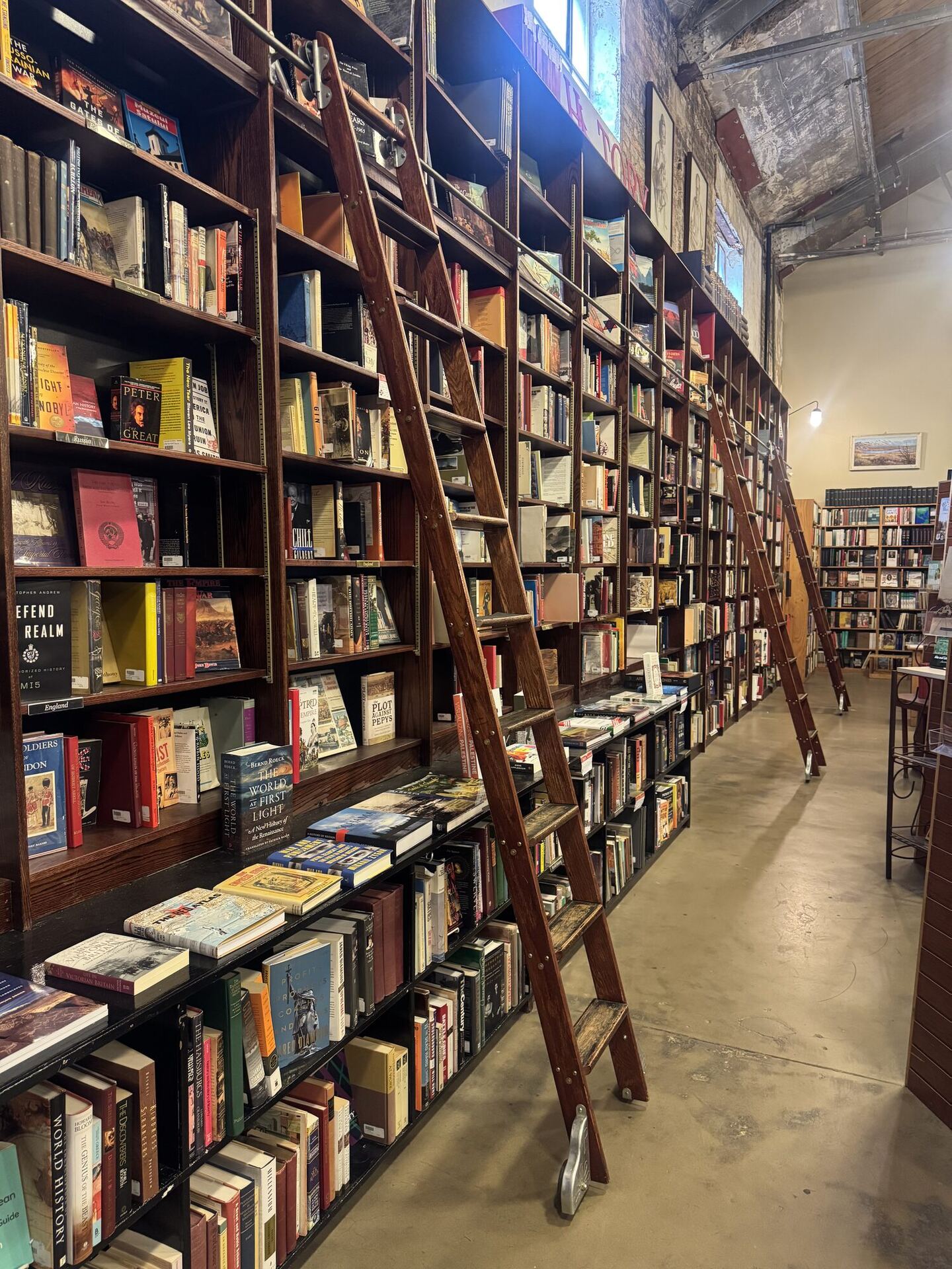 A long aisle of towering dark wooden bookshelves with a wooden rolling library ladder in the foreground and two more receding along the wall, under high windows, industrial pendant lights and exposed ductwork.