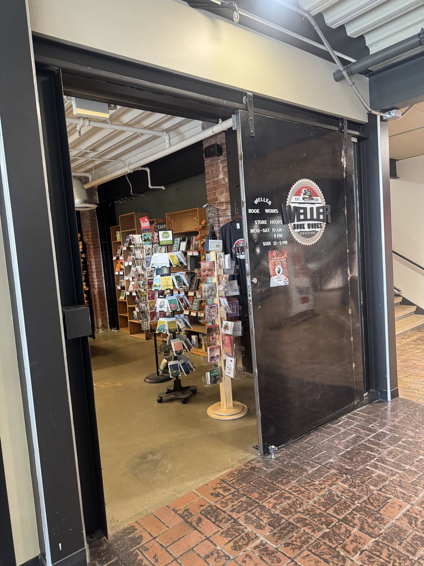 A black steel sliding door rolled open off a brick-paved corridor, painted with the round Weller Book Works seal and store hours, with spinning racks of greeting cards and shelves just inside.