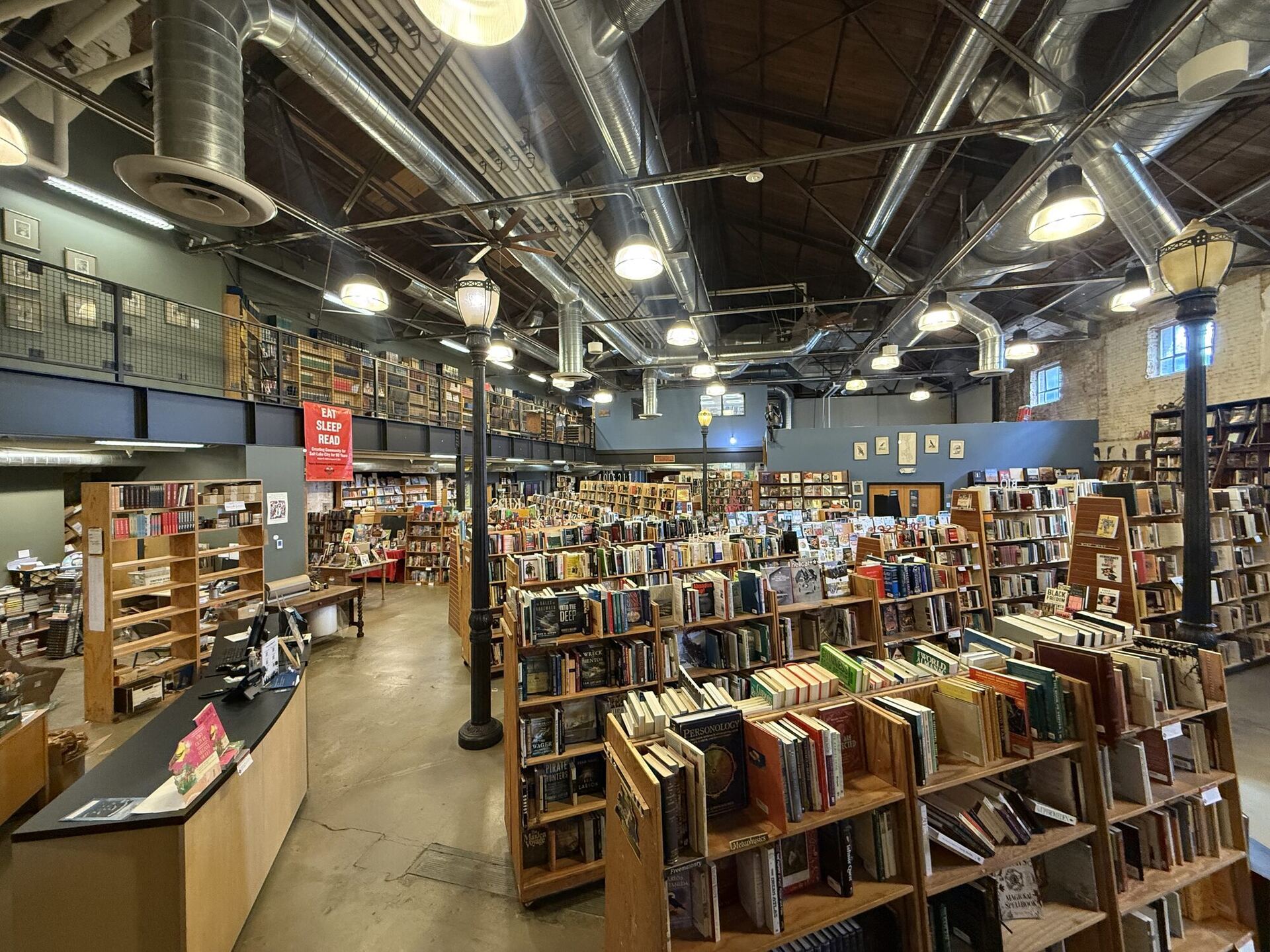 A wide, high-ceilinged warehouse bookshop with exposed wooden trusses, silver ductwork and hanging pendant lights, a curved front counter at left, a railed mezzanine lined with shelves, old-fashioned street lamps and long rows of wooden bookcases stretching back.