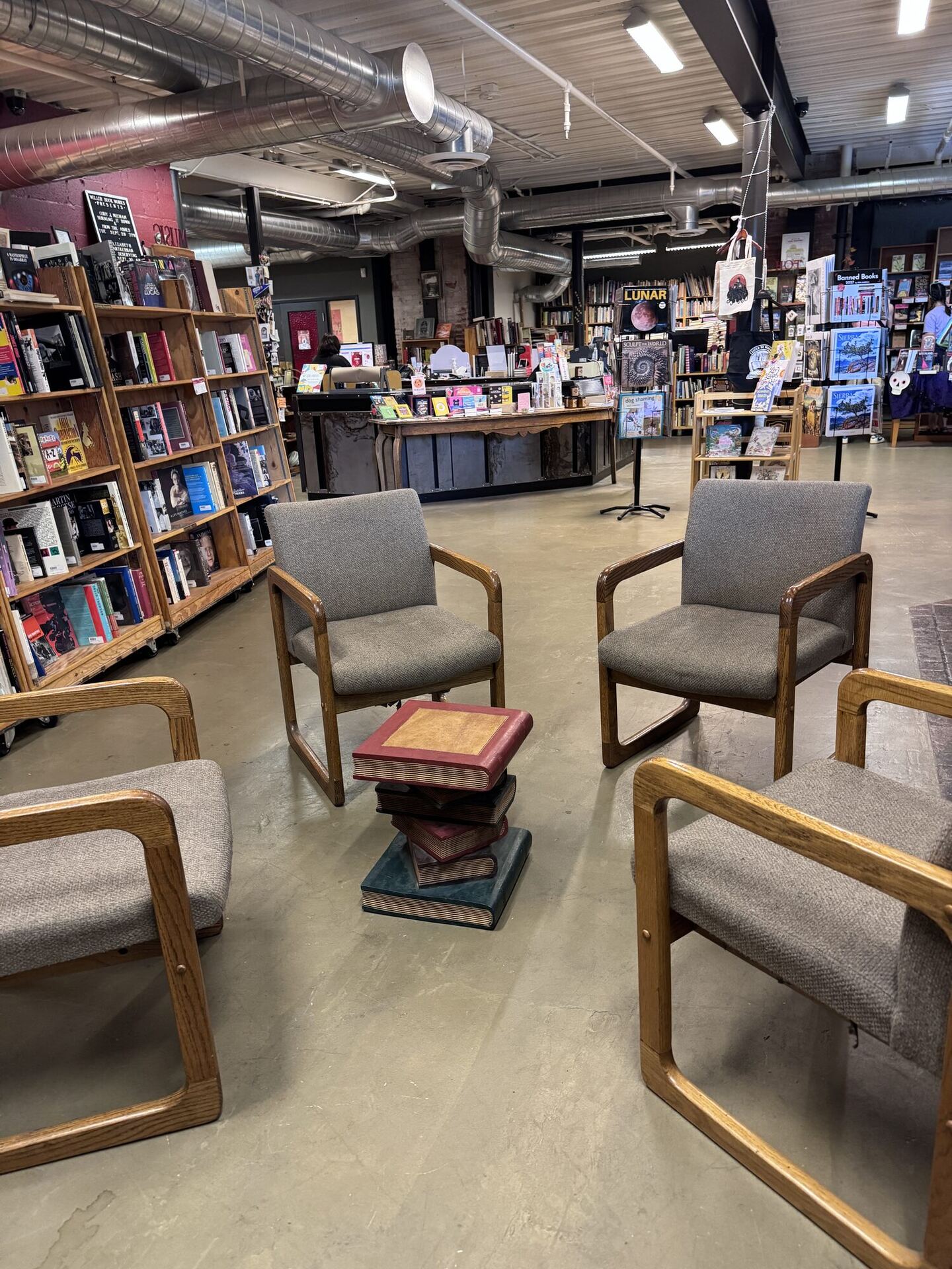 Four upholstered armchairs with oak frames arranged in a circle on a concrete floor around a small side table shaped like a stack of old books, with bookcases, the front counter and face-out displays behind.
