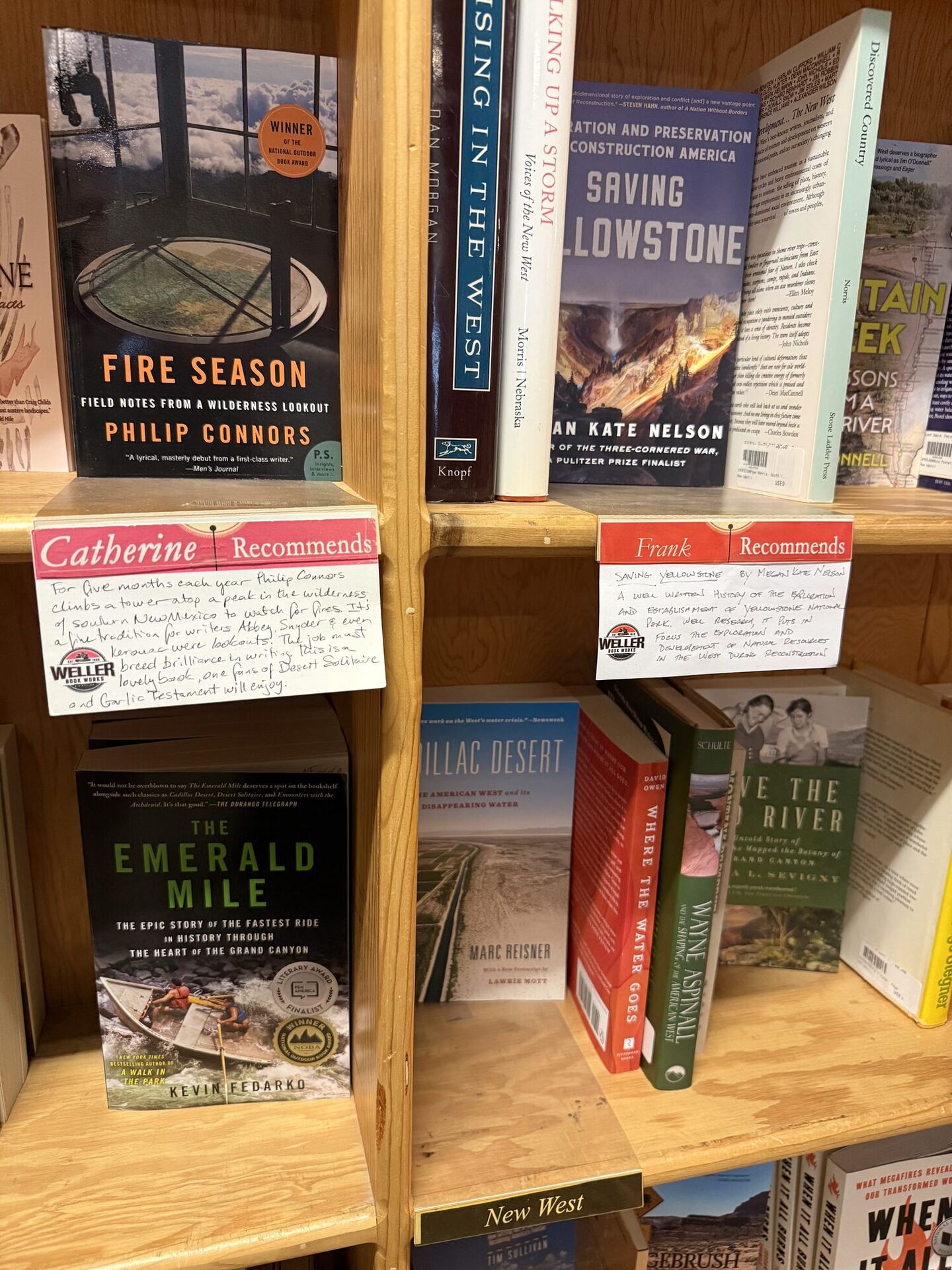 Two handwritten recommendation cards on a pine shelf of books about the American West. 'Catherine Recommends' sits under Fire Season by Philip Connors and 'Frank Recommends' under Saving Yellowstone, beside The Emerald Mile and Cadillac Desert.